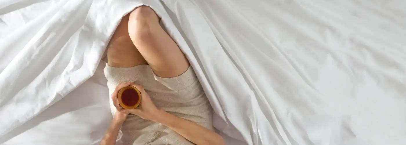 Overhead view of the legs of a person sitting in a pile of sheets and holding a cup of coffee