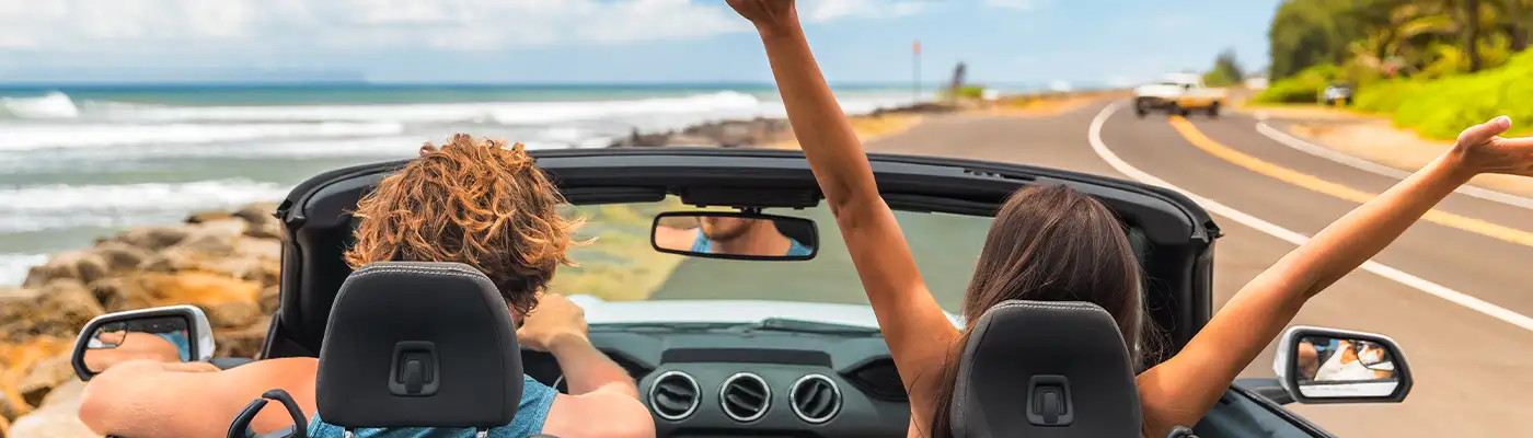Road trip summer vacation fun young couple driving convertible car on holiday travel destination banner panoramic. Woman with arms up happy, man driver having fun.